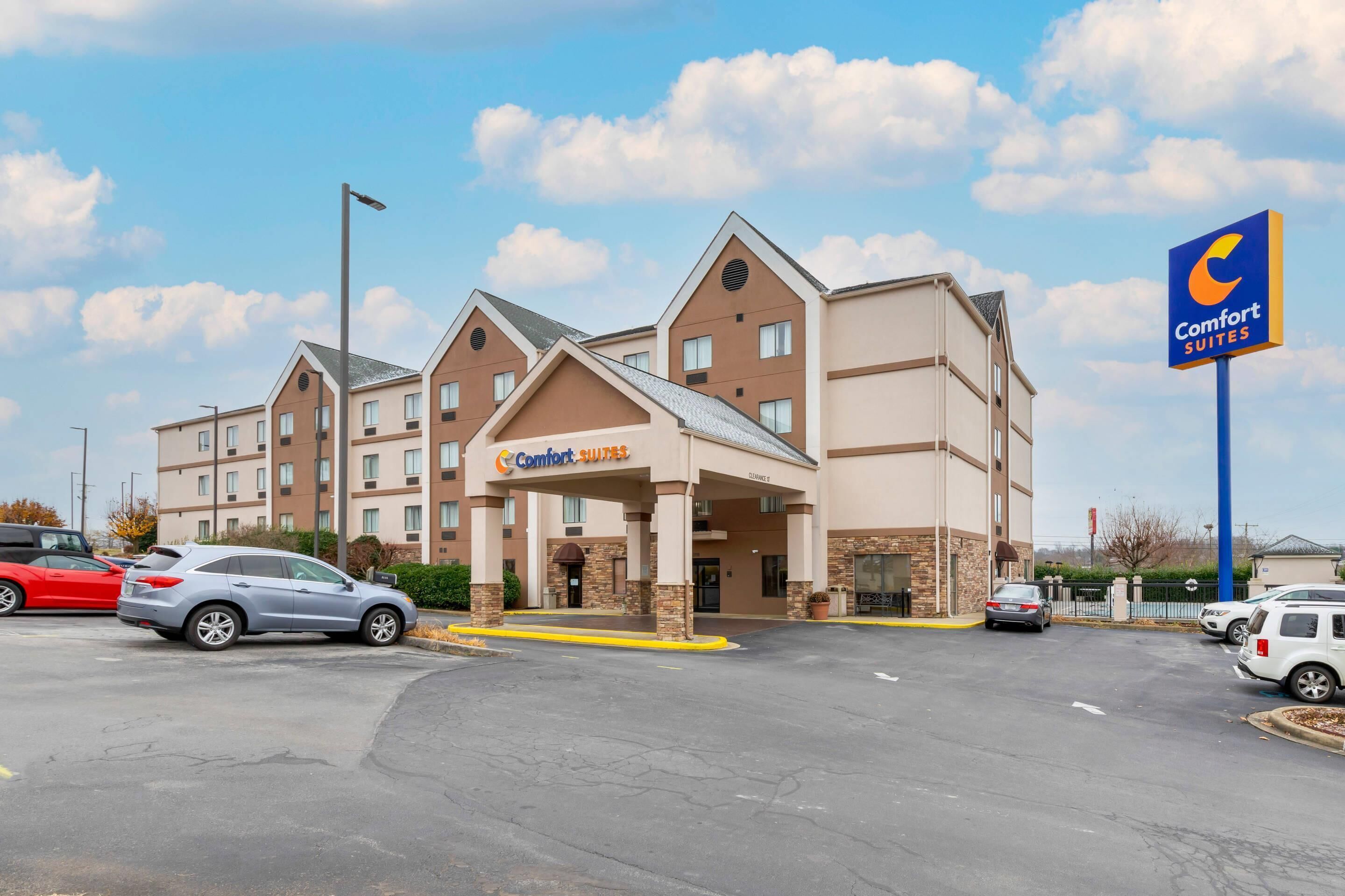 A Comfort Suites hotel with a modern design, featuring a central entrance, multiple floors, and a parking lot with several cars.