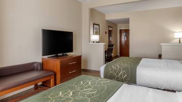 A hotel room with a television, a desk with drawers, a bench, two beds with green and white bedding, and a mirror on the wall.