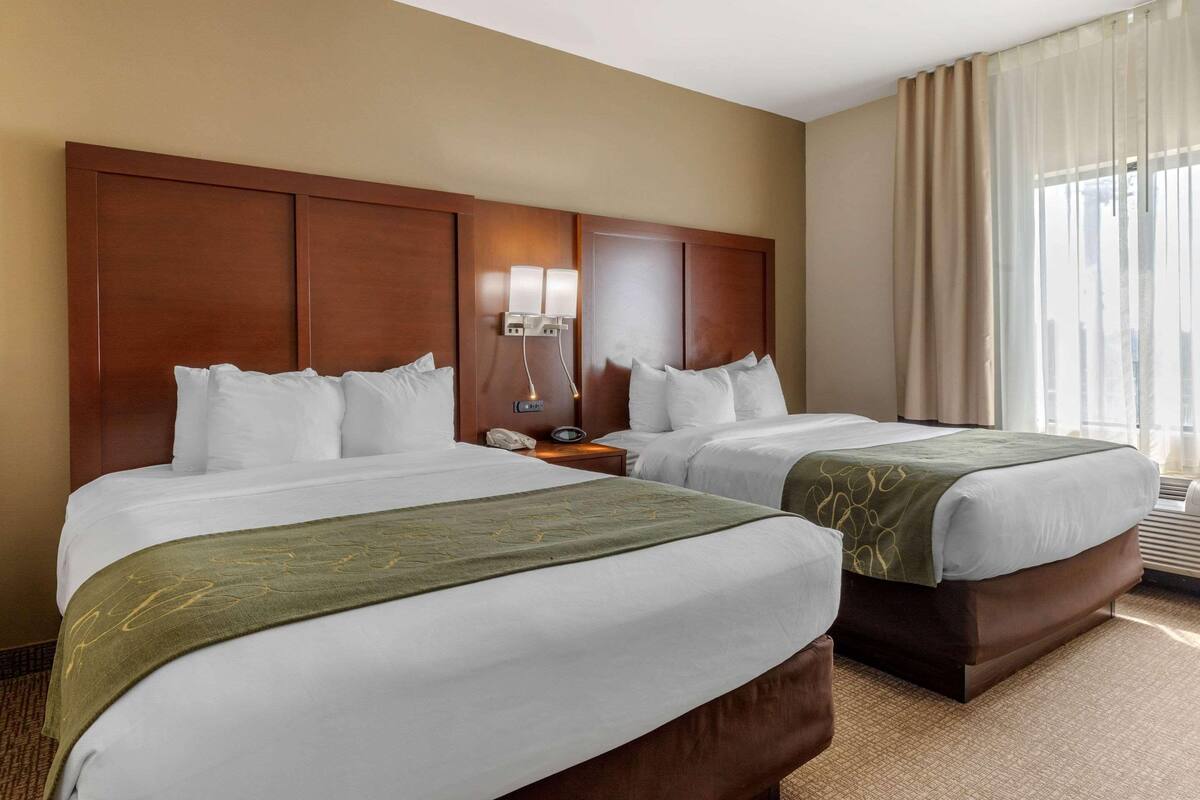 Habitación de hotel con dos camas, cabecera de madera, mesita de noche con lámpara y ventana con cortinas.