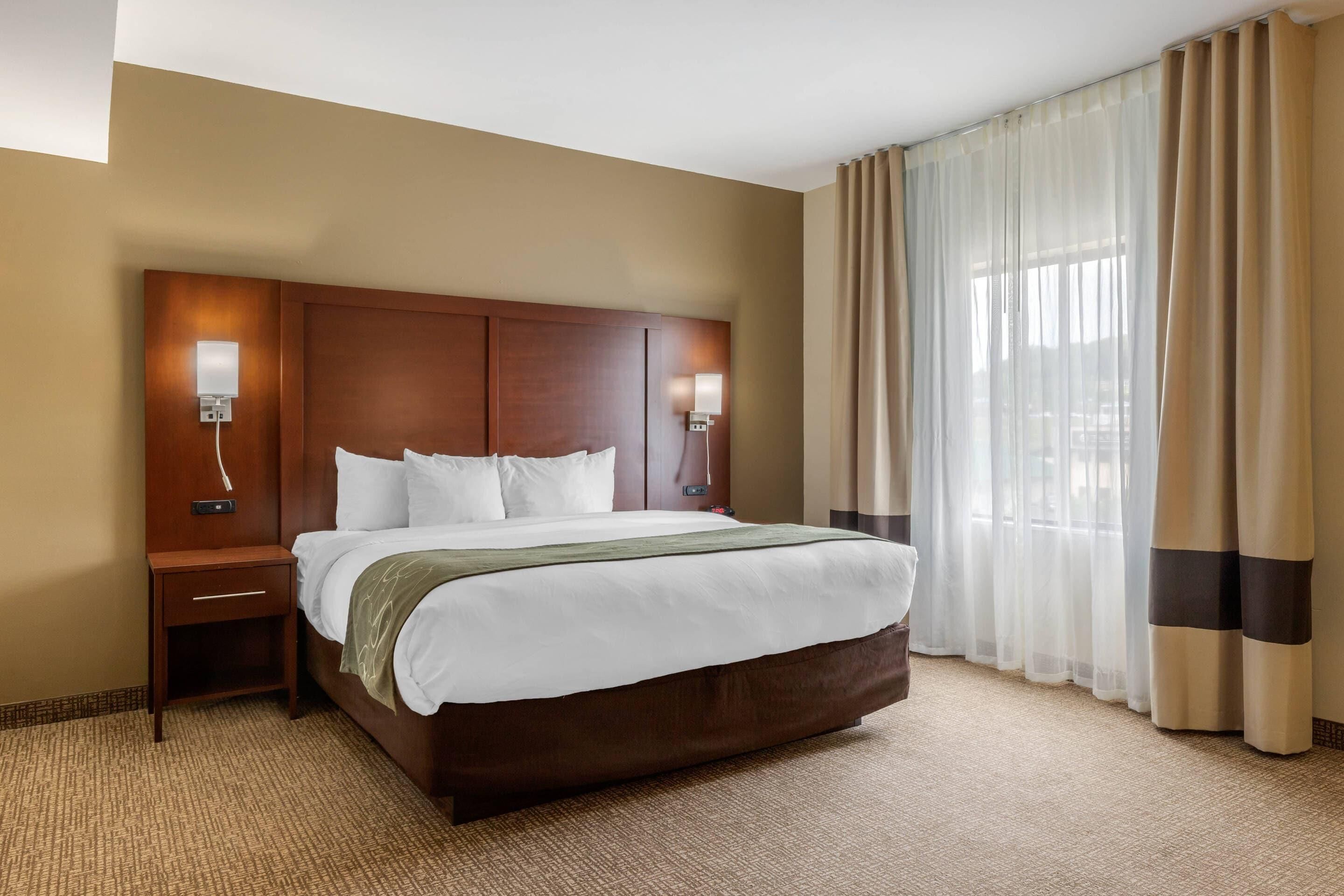 Een hotelkamer met een groot bed, twee nachtkastjes, een lamp aan de muur en een raam met dunne gordijnen.