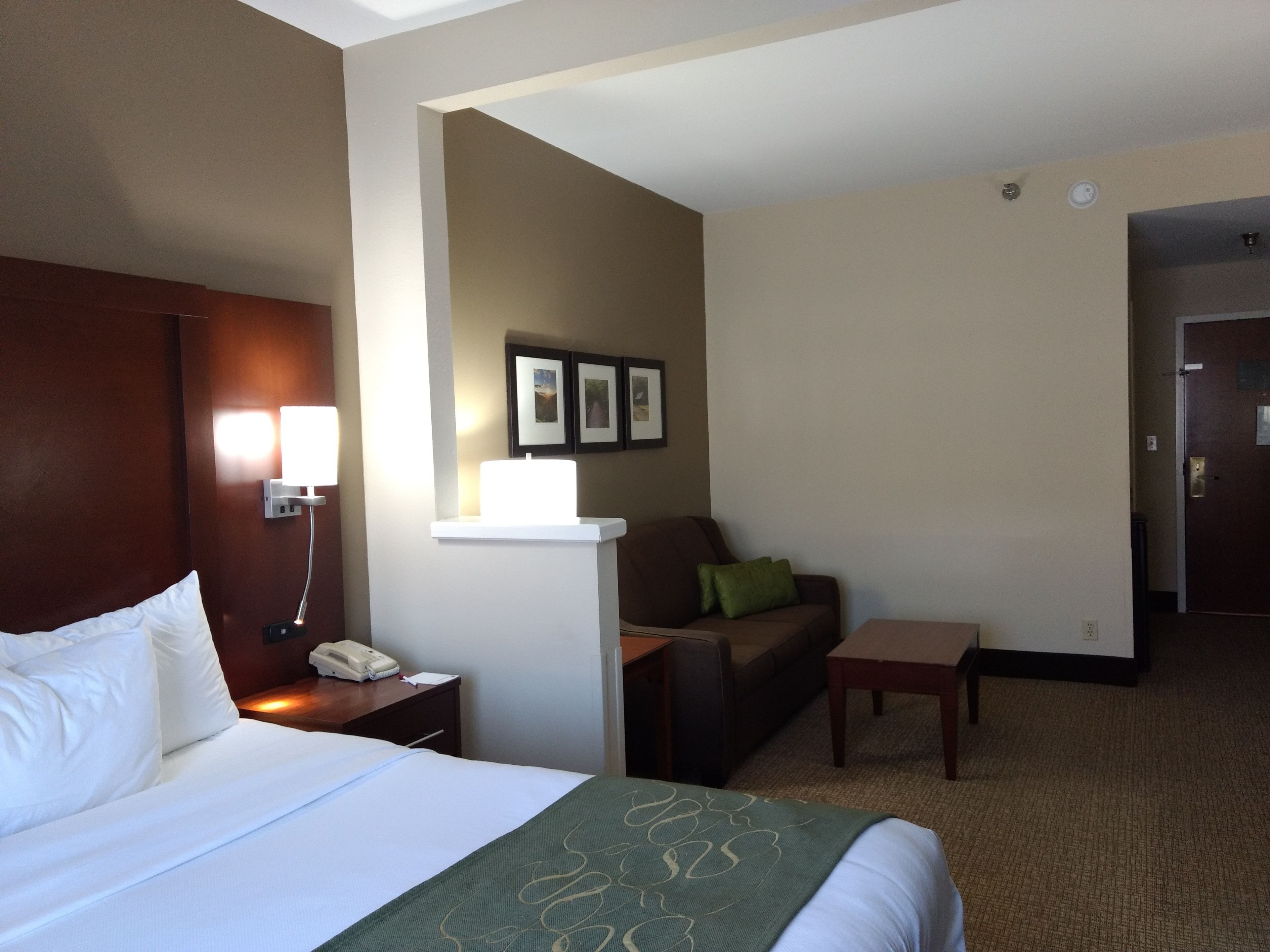 Habitación de hotel con cama, escritorio, silla, sofá y una mesita. Hay un teléfono de pared y una lámpara sobre el escritorio.