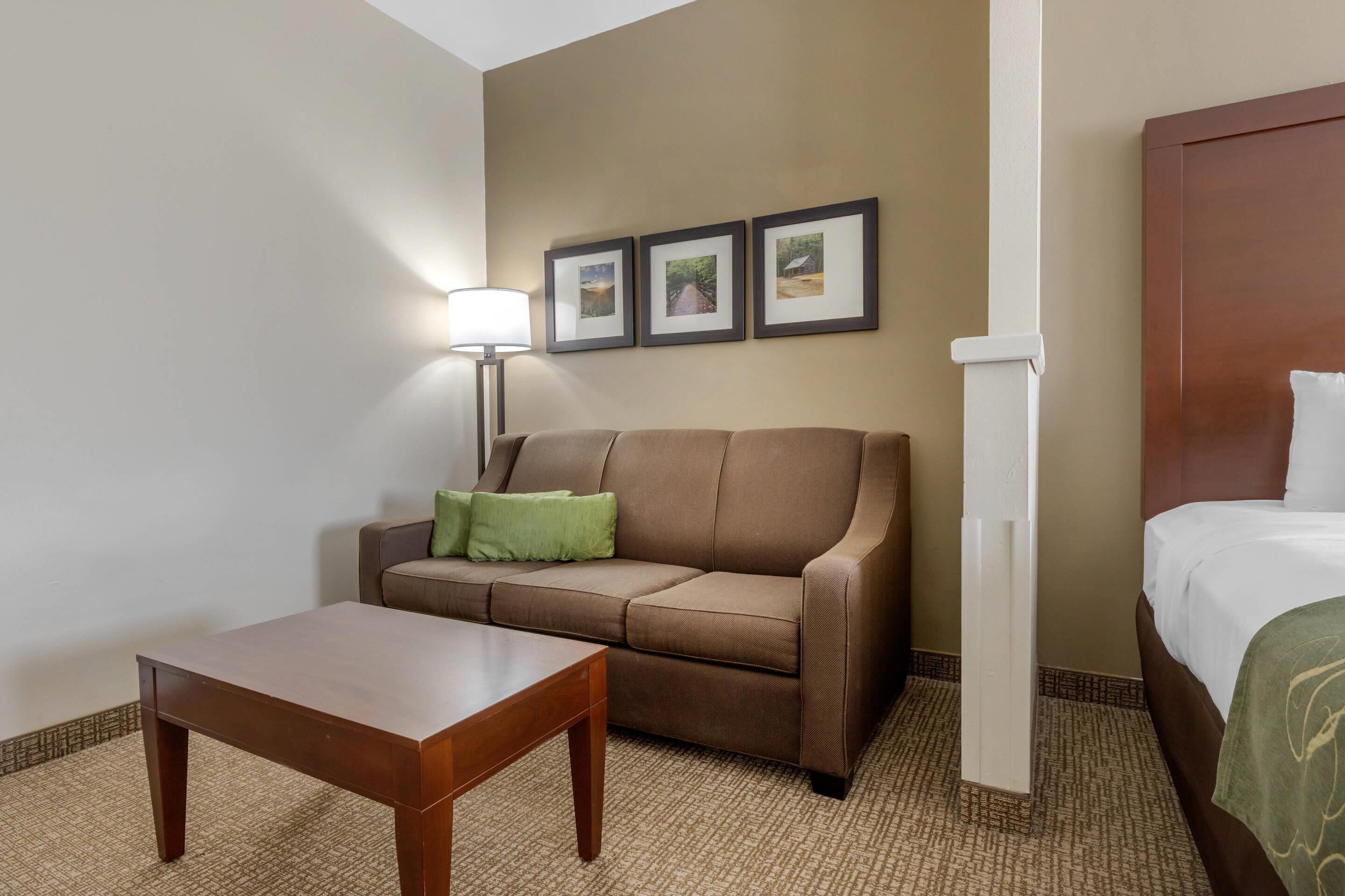 A hotel room with a brown sofa, a wooden table, a bed with white bedding, a lamp, and three framed pictures on the wall.