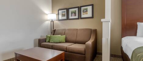 A hotel room with a brown sofa, a wooden table, a bed with white bedding, a lamp, and three framed pictures on the wall.