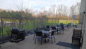 An outdoor patio area with tables and chairs, a grill, and a view of a sports field.