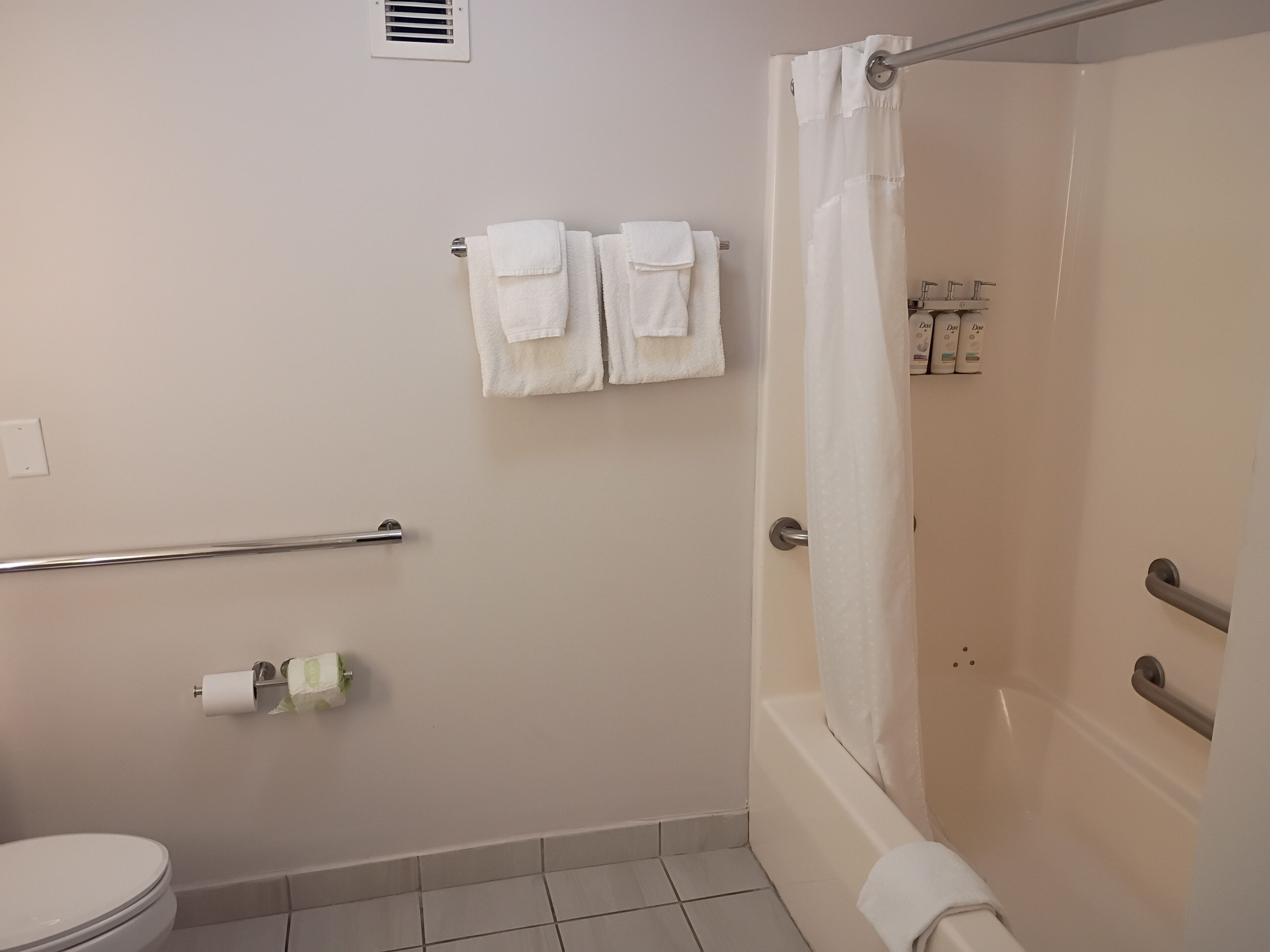 Un baño con inodoro, ducha con cortina, barra de apoyo y dos toallas en un perchero.