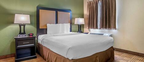 A hotel room with a bed, two bedside tables with lamps, a nightstand, a window with curtains, and a wall clock.