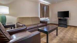 A hotel room with a brown leather sofa, a small black table, a television on a stand, a ceiling fan, and a window with curtains. - Extended Stay America Select Suites Denver Tech Center South (Englewood)