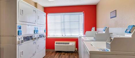 A laundry room with multiple washing machines, a dryer, and a window with blinds.