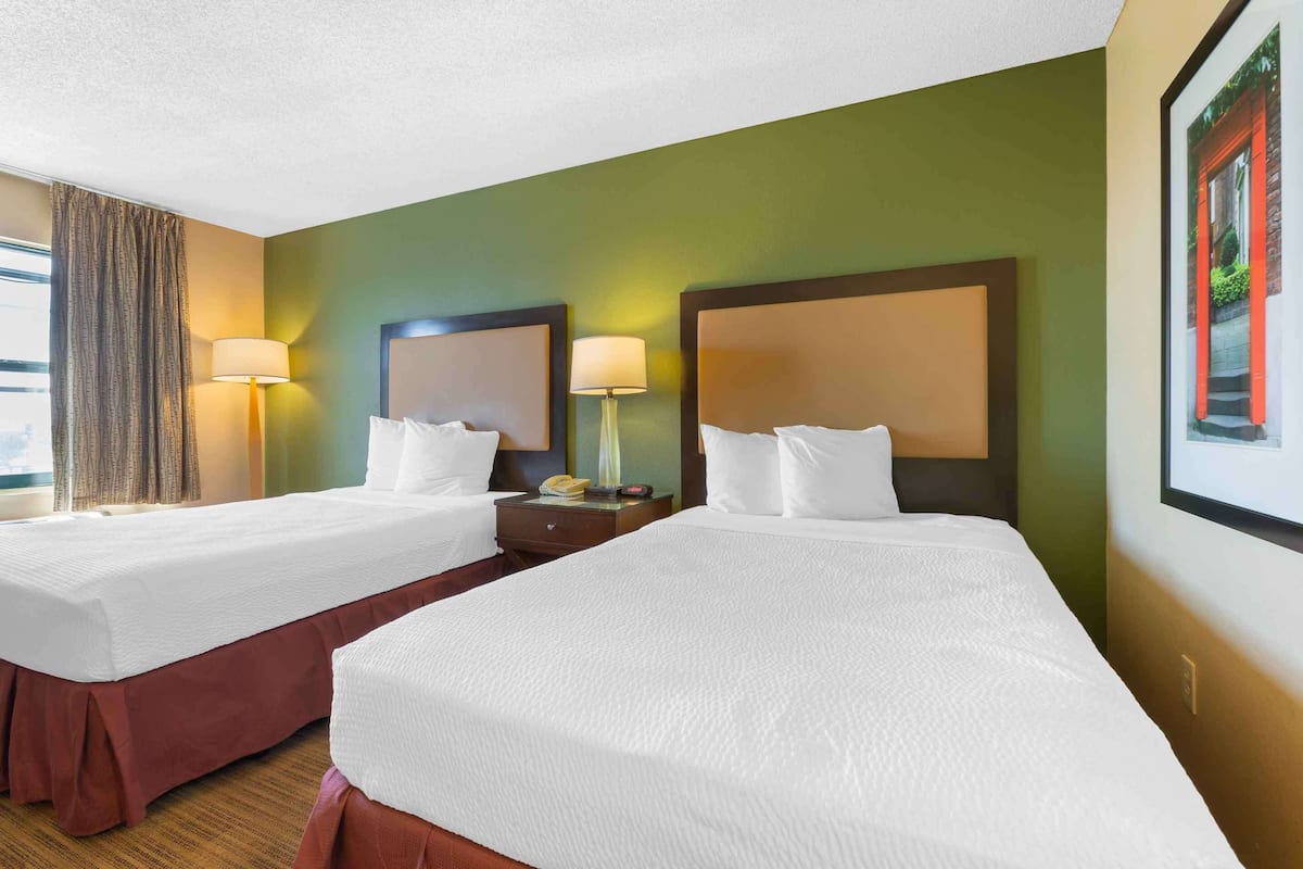 A hotel room with two beds, a nightstand, a lamp, and a window with curtains.