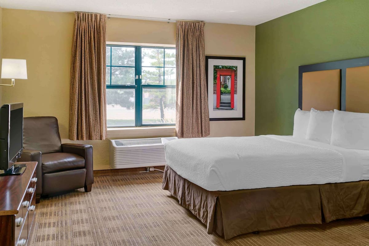 A hotel room with a large bed, a desk with a chair, a window with curtains, and a framed picture on the wall.