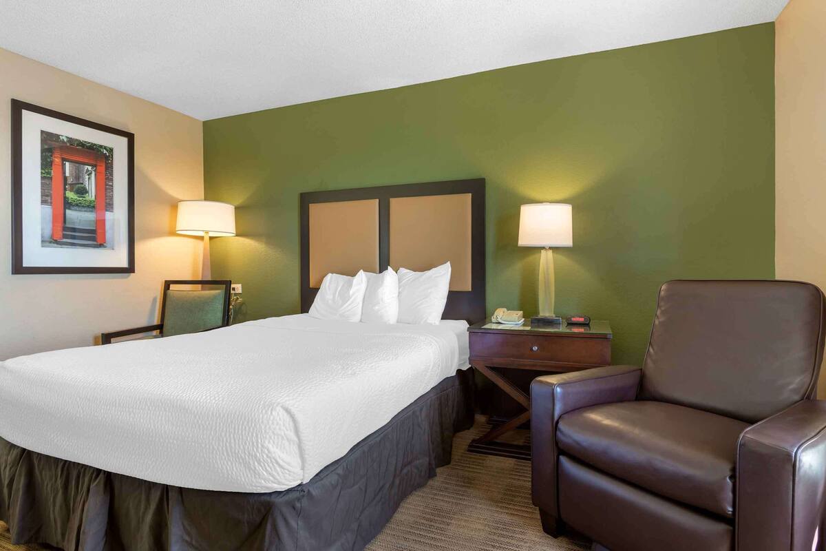 A hotel room with a bed, two bedside lamps, a chair, a nightstand, and a framed picture on the wall.