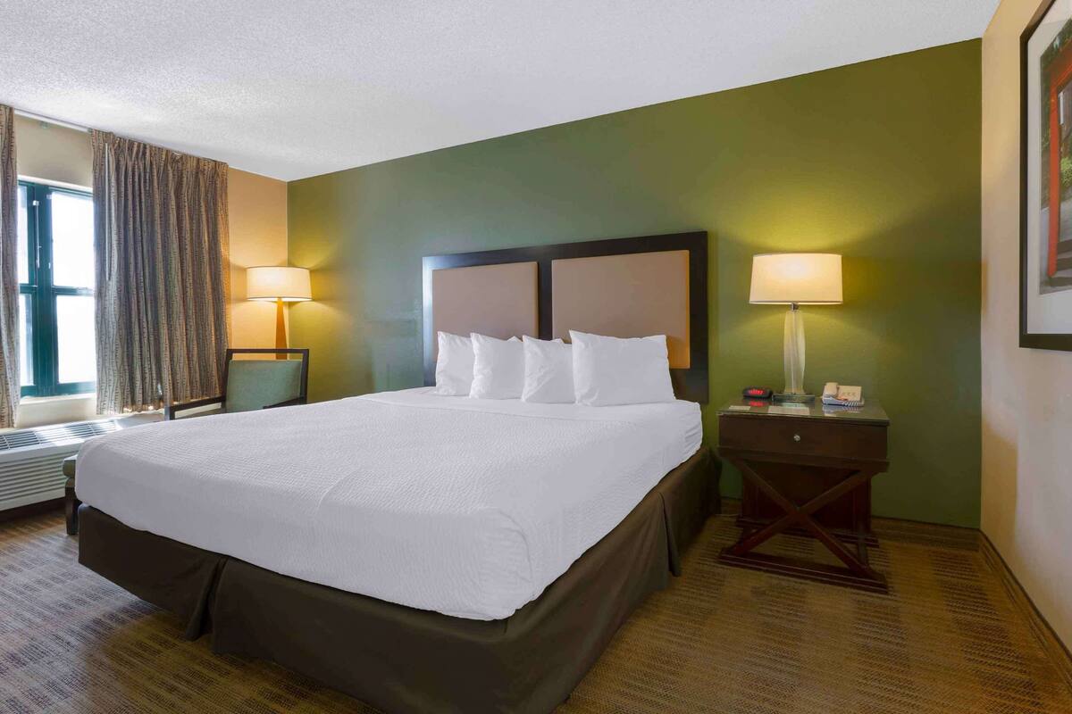 A hotel room with a large bed, two bedside tables with lamps, a window with curtains, and a framed picture on the wall.