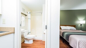 A room with a wooden floor, a white toilet, a shower, and a bed with a green and pink bedspread. - Studio 51 Extended Stay Hotel (Phoenix)