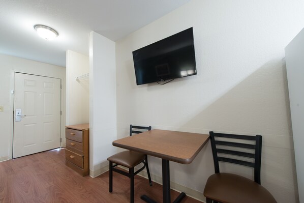 Una habitación pequeña con una mesa de madera y dos sillas, un televisor montado en la pared y una puerta.
