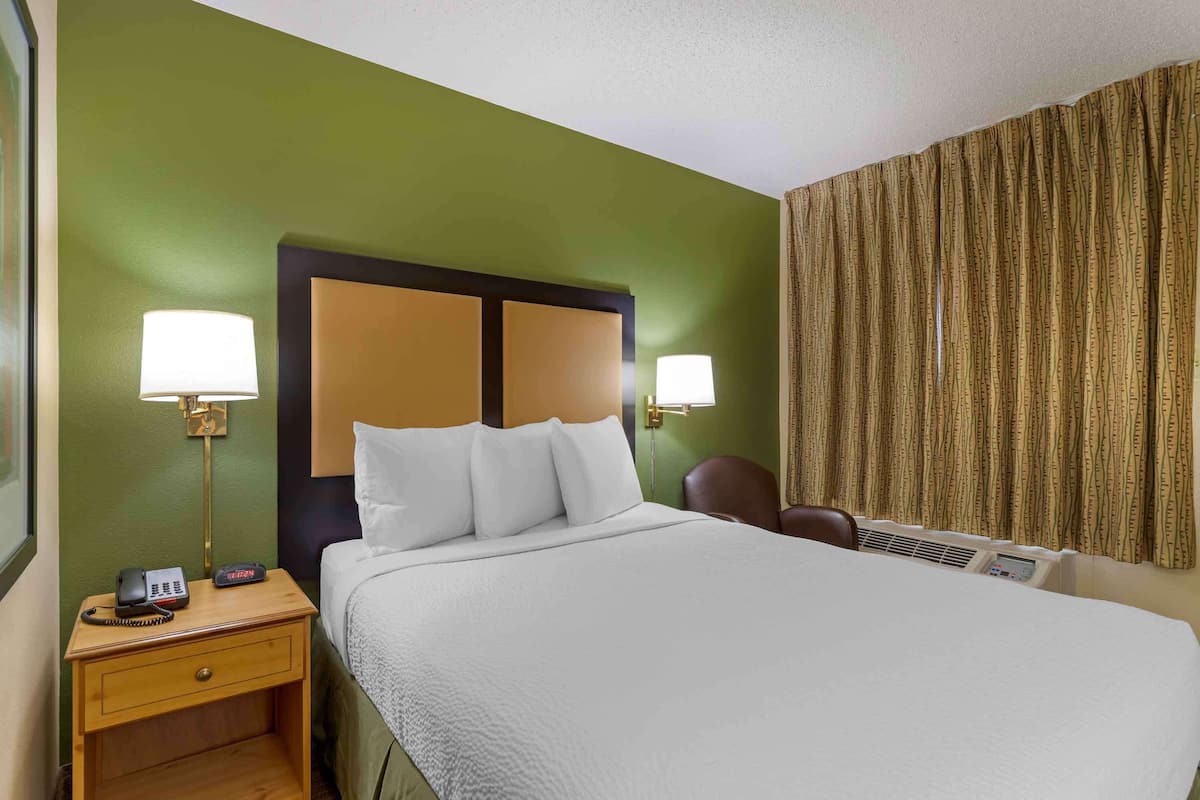 A hotel room with a bed, bedside lamps, a nightstand with a phone, a chair, and a window with curtains.
