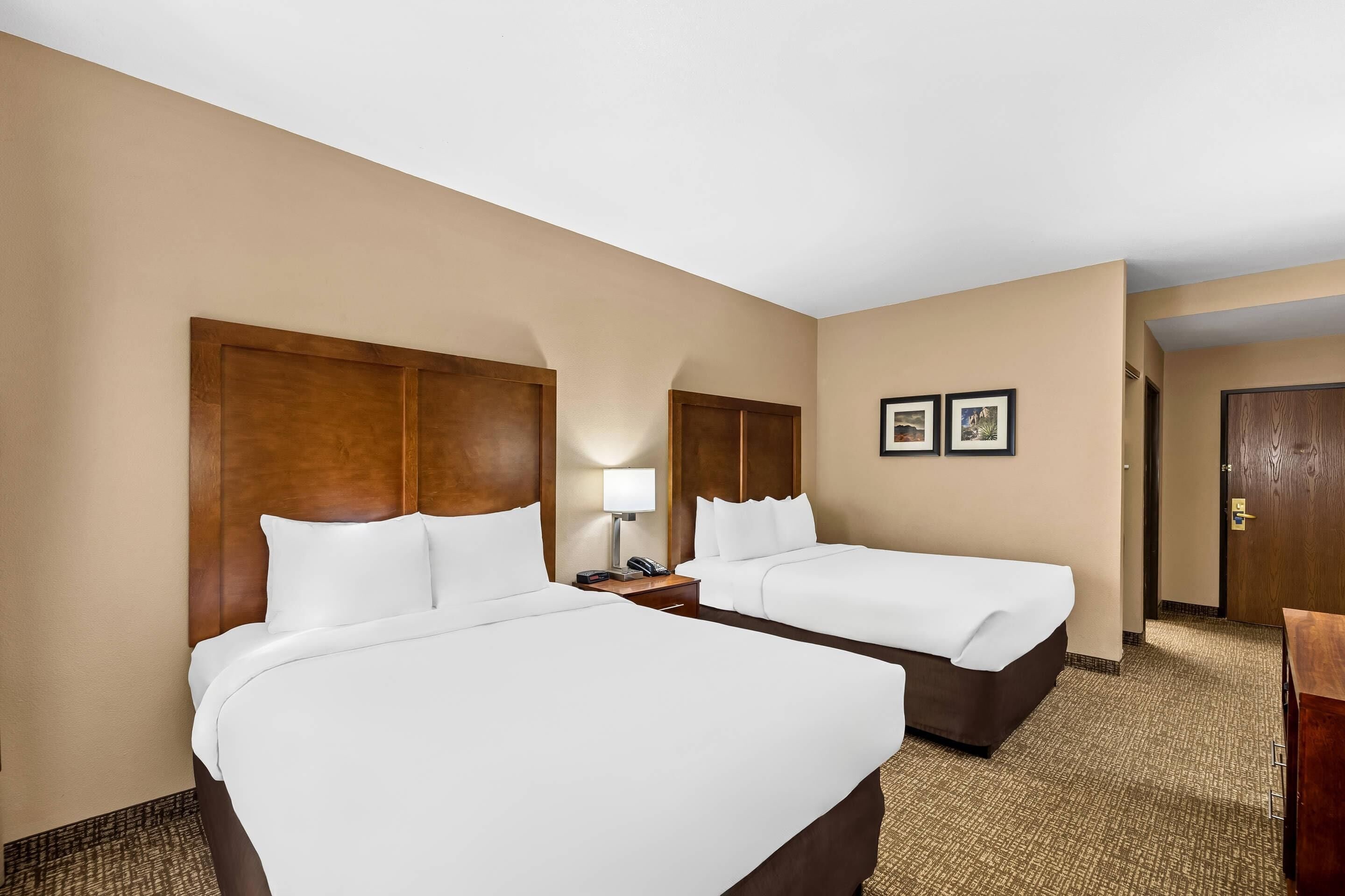 A hotel room with two beds, a nightstand with a lamp, and a door leading to another room.
