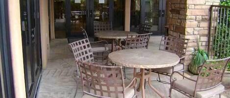 Un patio al aire libre con mesas redondas y sillas de mimbre, rodeado por un muro de piedra y puertas de vidrio que dan al interior.