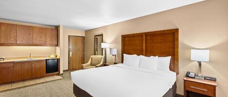 A hotel room with a large bed, a desk with a lamp, a chair, a minibar, and a bathroom door.