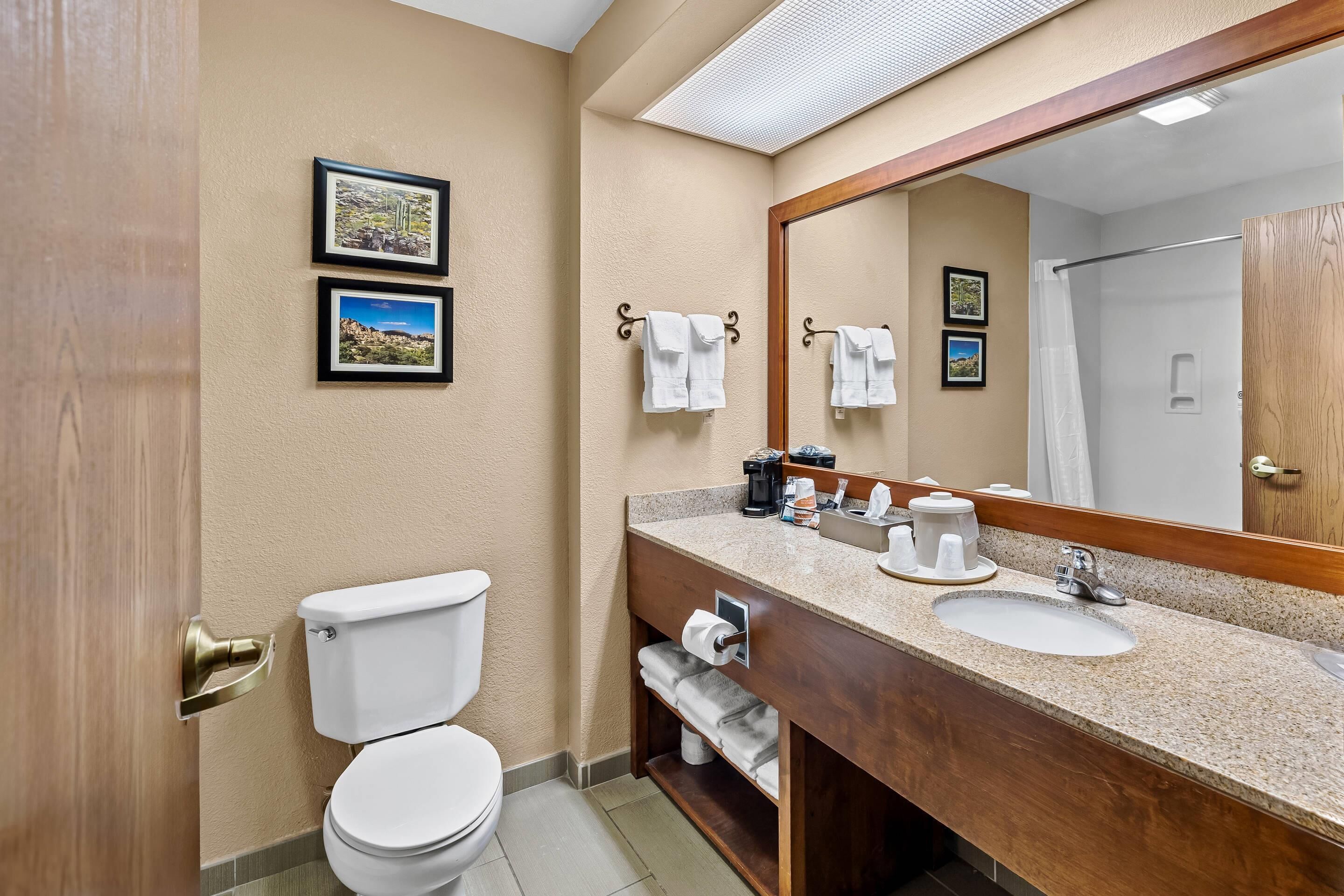 A hotel bathroom with a toilet, sink, and mirror. There are towels hanging on hooks and a shower curtain visible in the reflection.