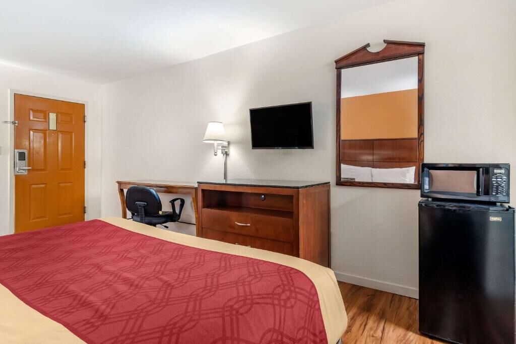 Una habitación de hotel con cama, escritorio, silla, televisor, espejo y refrigerador.