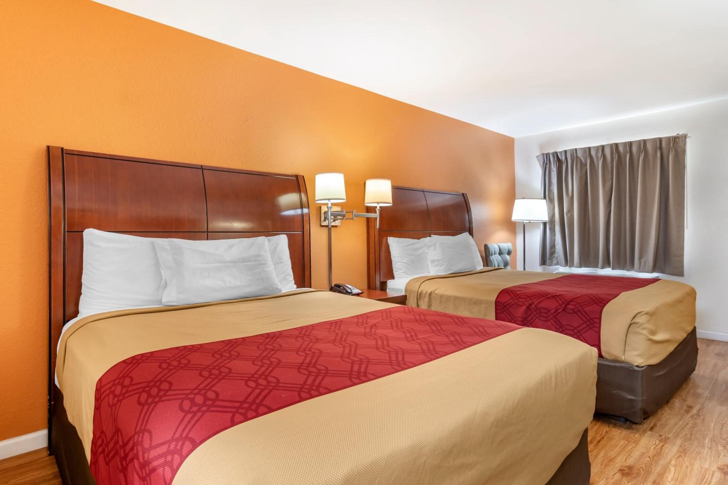 Habitación de hotel con dos camas, cada una con cabecero de madera, colcha estampada roja y manta color beige. Hay lámparas de noche y mesitas de noche con teléfono.
