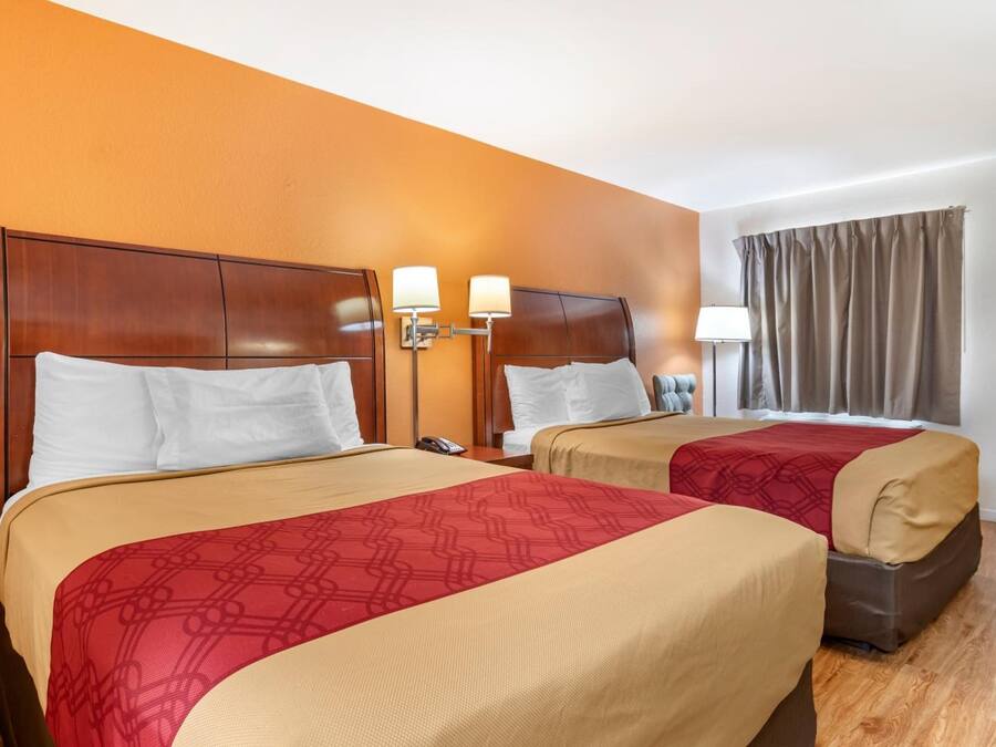 Ein Hotelzimmer mit zwei Betten, jeweils mit einem hölzernen Kopfteil, einer rot gemusterten Bettdecke und einer beigen Decke. An den Betten befinden sich Nachttischlampen und ein Nachttisch mit einem Telefon.