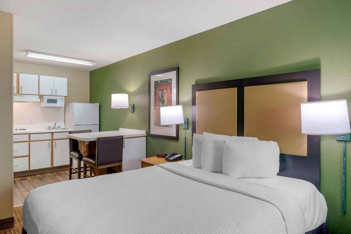 Hotelkamer met een bed, een bureau, een stoel, een kleine kitchenette en een nachtkastje met lamp.