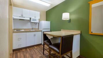 Una cocina compacta con armarios blancos, microondas, refrigerador y una pequeña mesa de comedor con una silla.