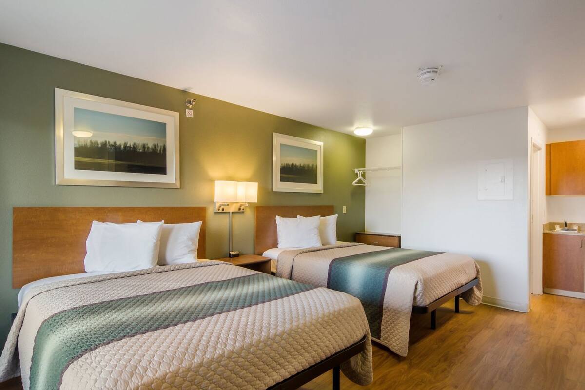 A hotel room with two beds, a nightstand, a lamp, a painting, and a small kitchenette.