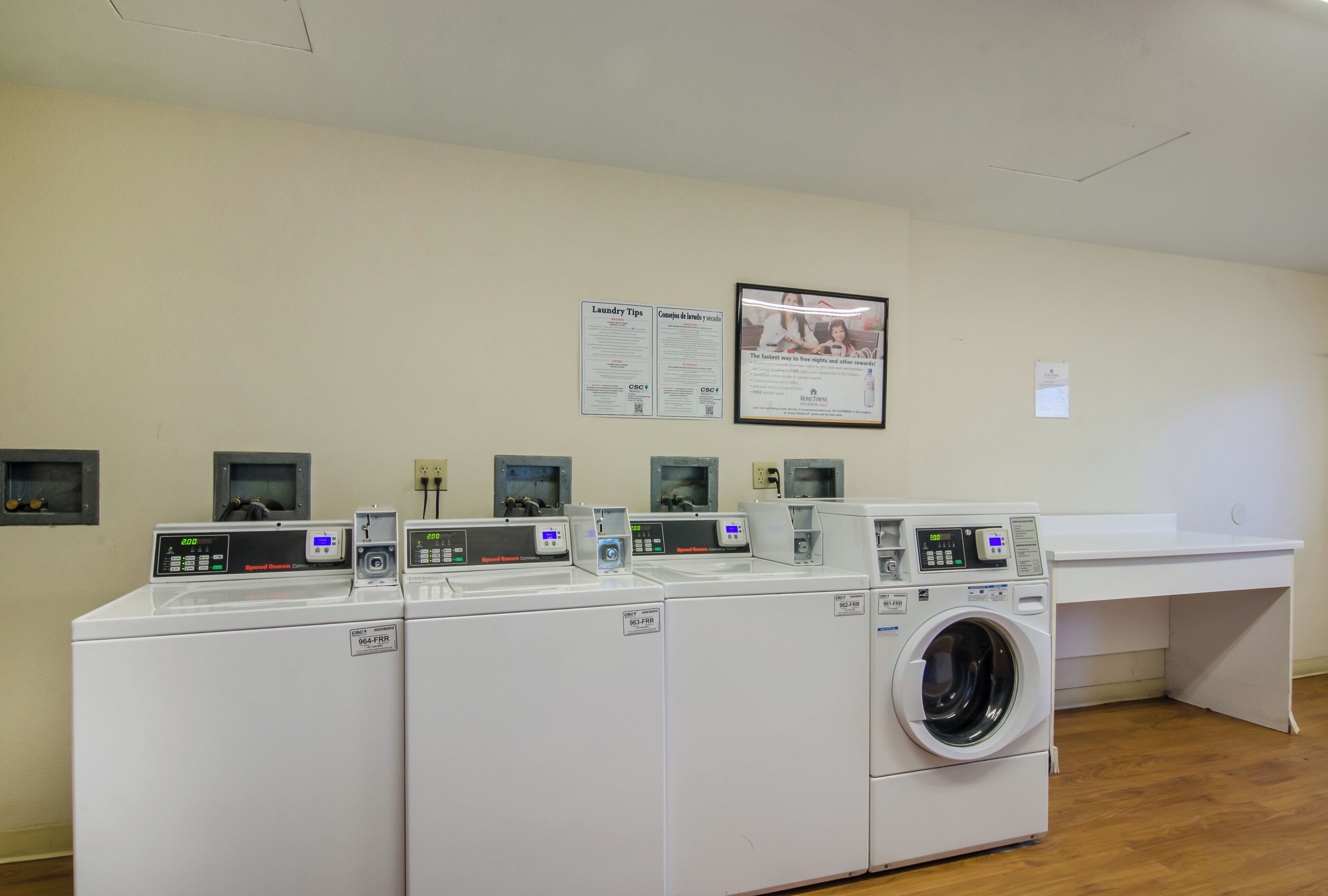 A laundry facility with multiple washing machines and dryers, a wall-mounted sign with laundry tips, and a framed advertisement.