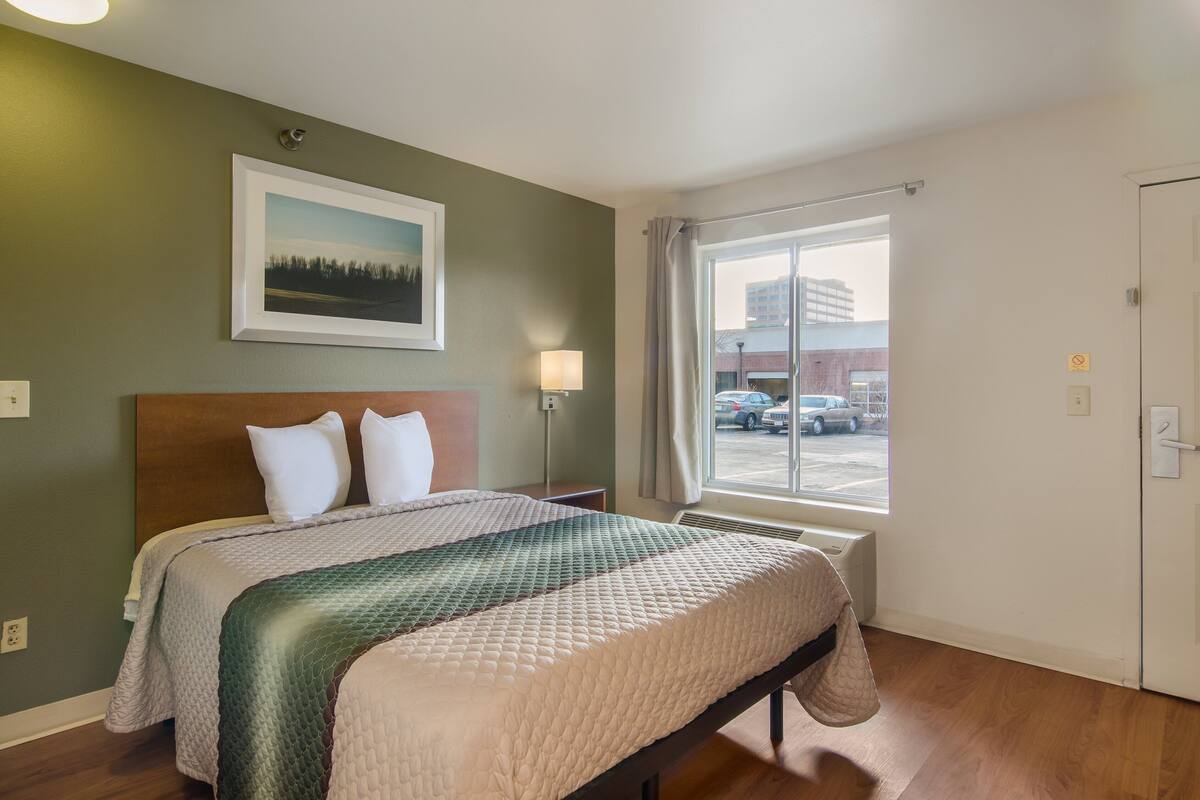 A hotel room with a bed, a window with a view of the parking lot, and a painting on the wall.