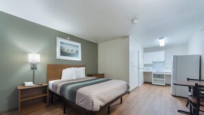 Una camera d'albergo con un letto, comodino, lampada, un quadro appeso e un angolo cottura con microonde e frigorifero.