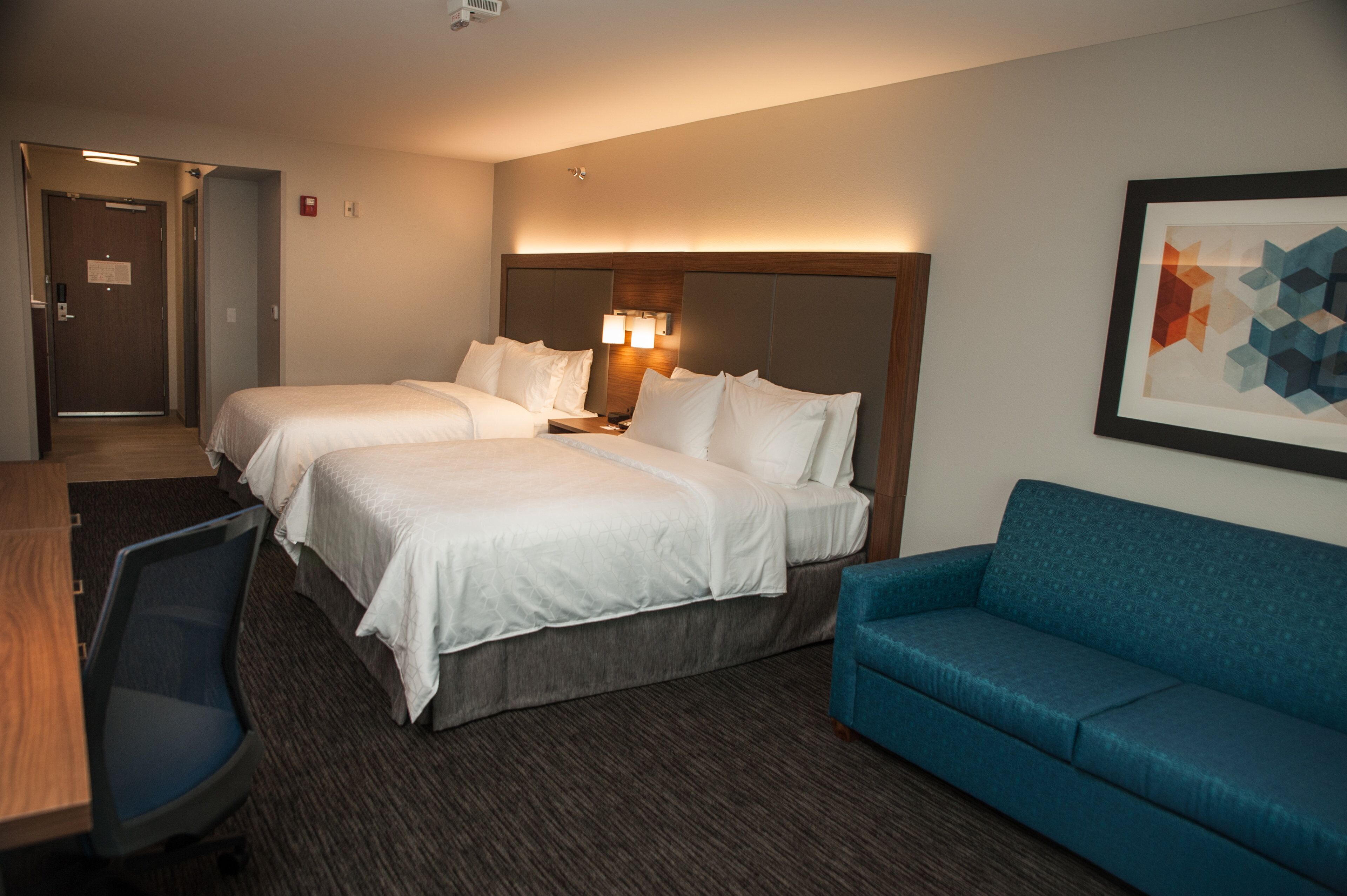 A hotel room with two beds, a desk, a blue sofa, and a framed abstract art piece on the wall.