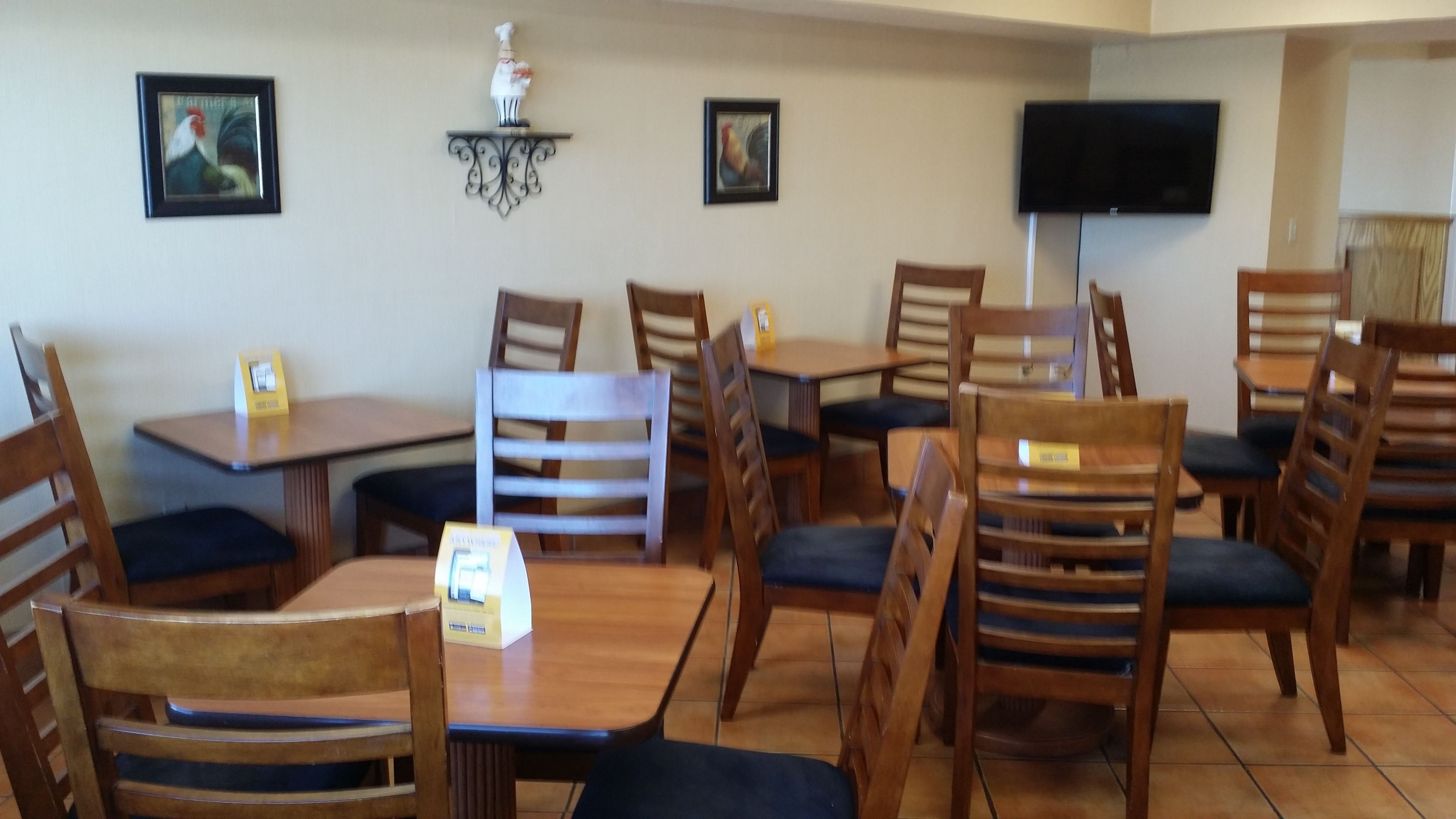 Un área de comedor con mesas y sillas de madera, un televisor en la pared y obras de arte enmarcadas sobre las mesas.