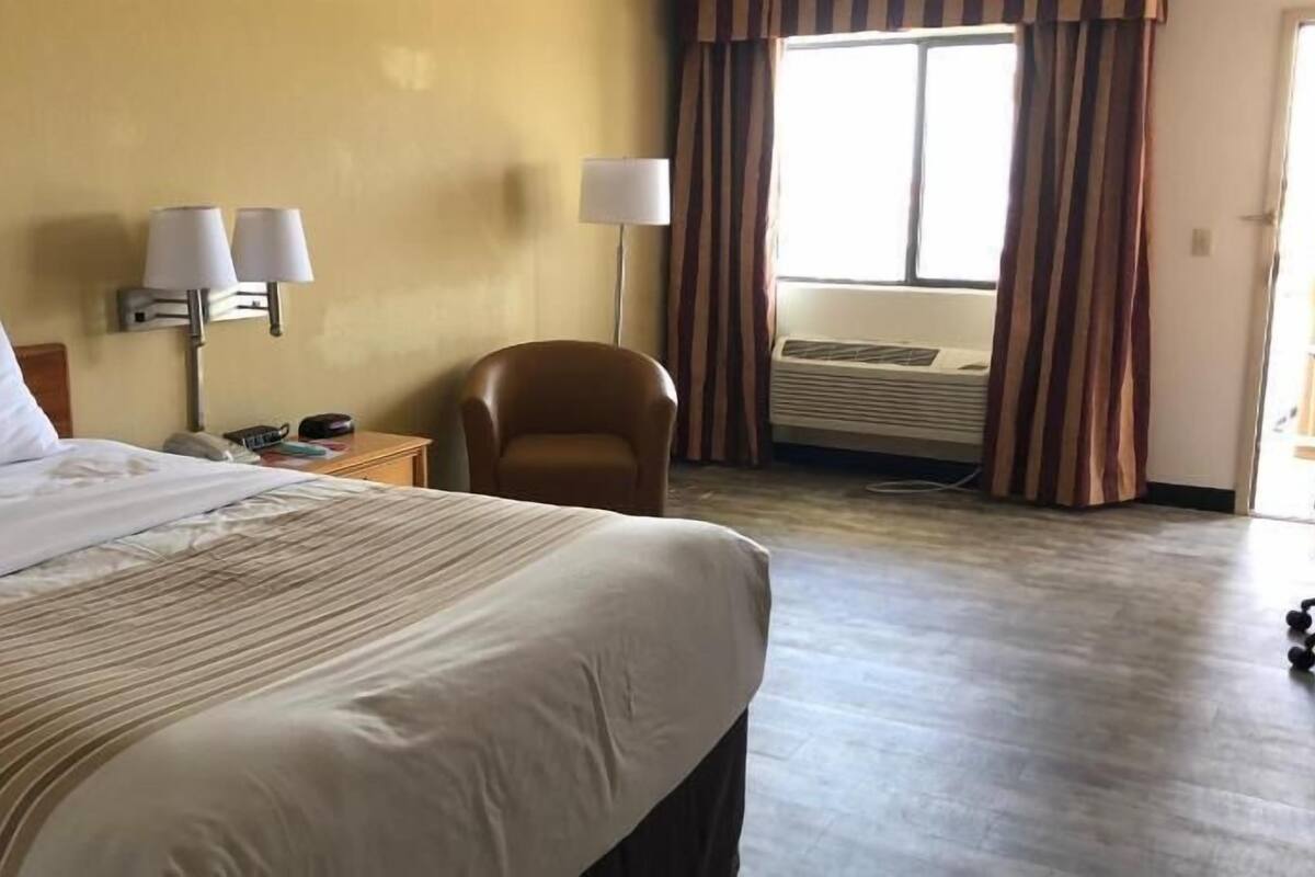 Una habitación de hotel con una cama grande, una silla, un escritorio con lámpara y un equipo de aire acondicionado.