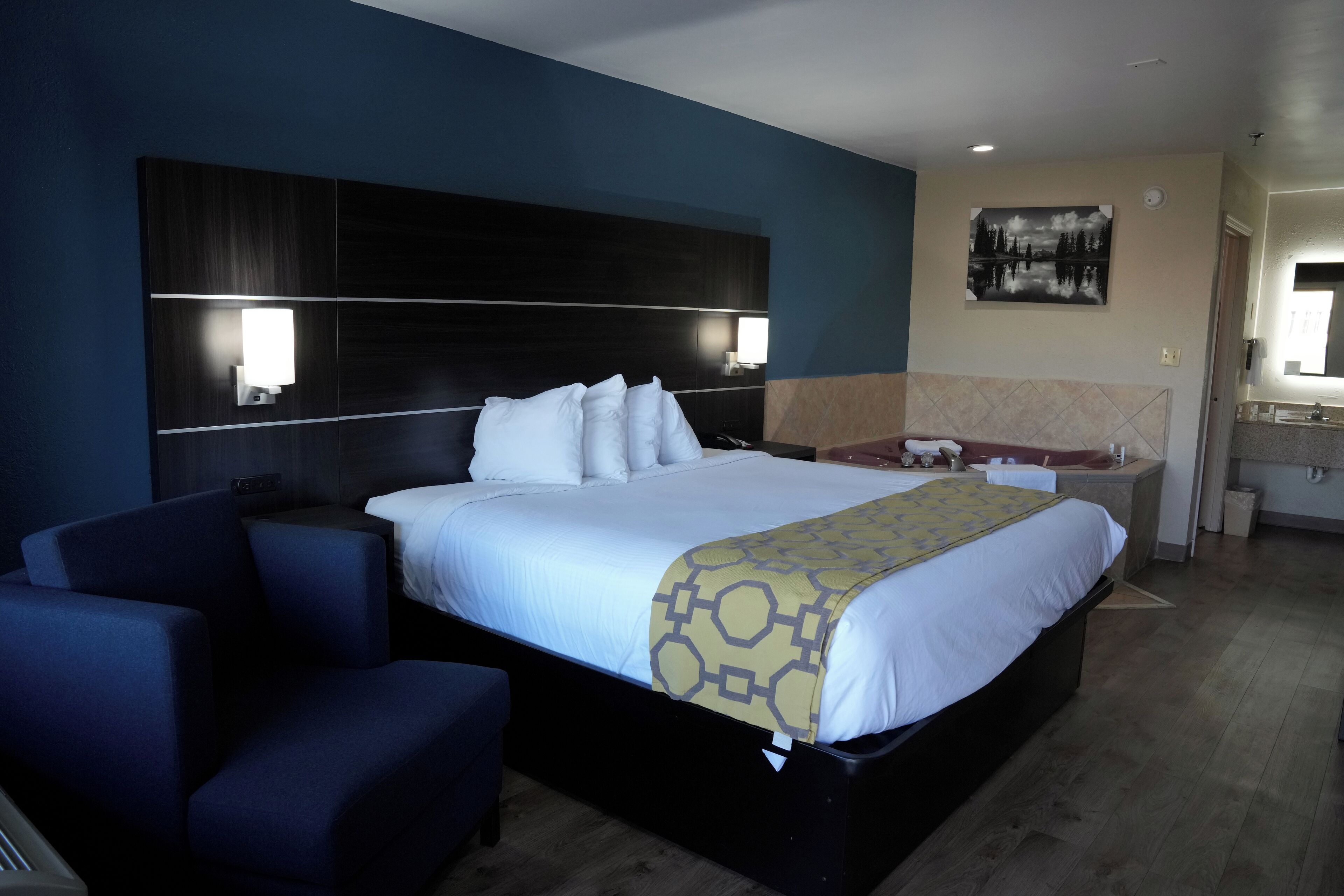 A hotel room with a large bed, a bedside table, a chair, a desk, a mirror, and a bathroom with a sink and toilet.