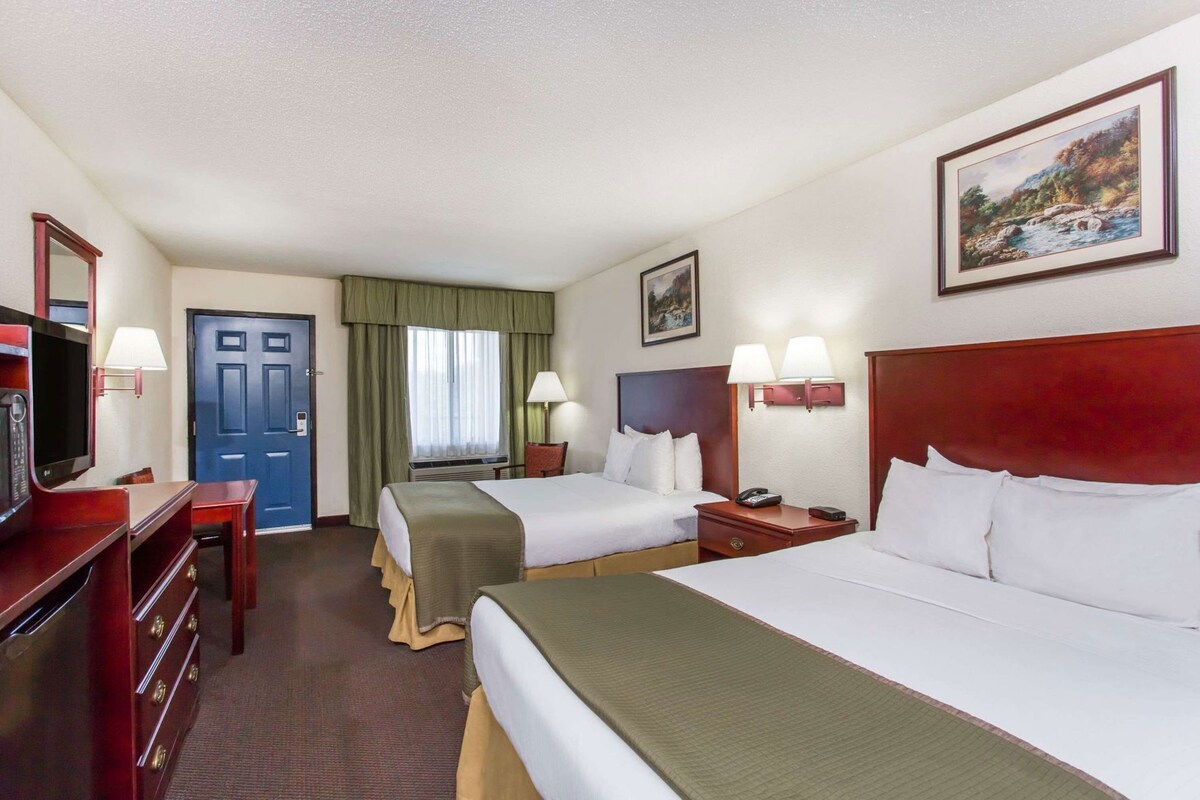 A hotel room with two beds, a desk with a TV, a door, and a painting on the wall.