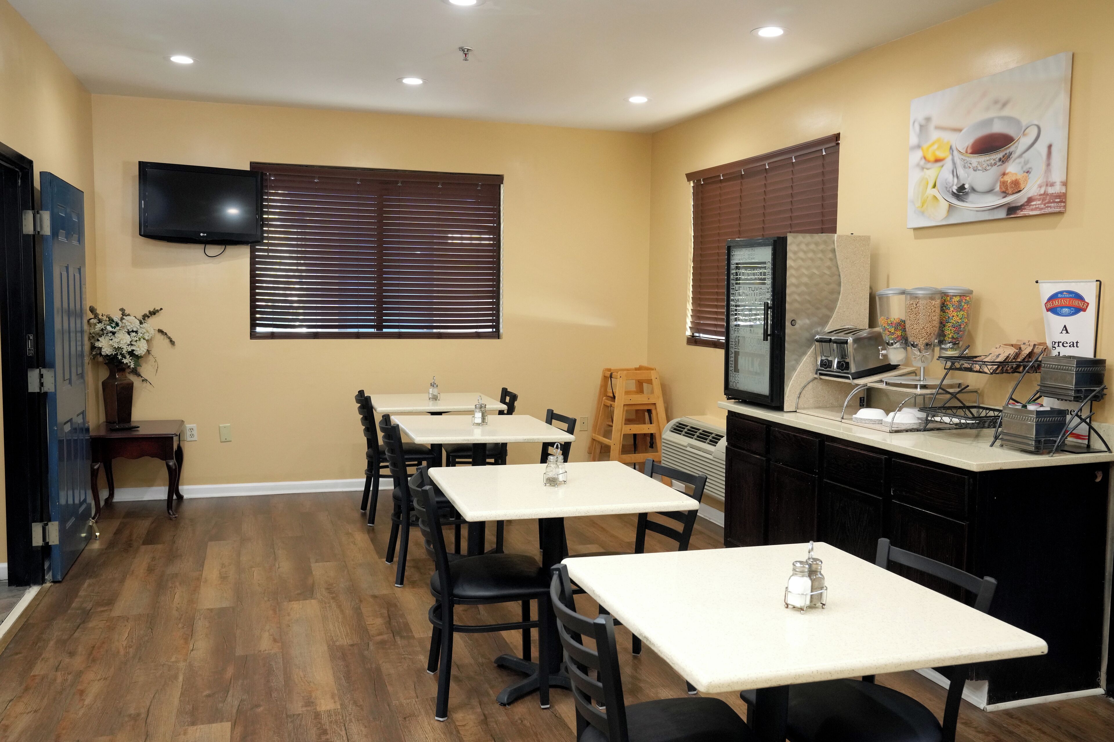 Un comedor con mesas y sillas, un televisor montado en la pared y una cocina con electrodomésticos y una estación de café.