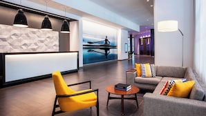 A modern lobby with a reception desk, a yellow armchair, a grey sofa with colorful cushions, and a large artwork depicting a person rowing a boat. - Le Méridien Boston Cambridge (Cambridge)