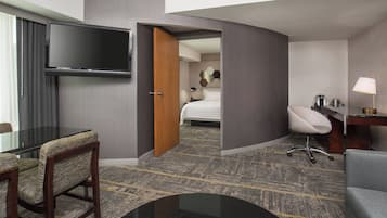 Une chambre d’hôtel dotée d’un téléviseur à écran plat fixé au mur, d’un lit avec une tête de lit à motifs géométriques, d’un bureau avec une lampe et d’un coin salon comprenant une chaise et un canapé.
