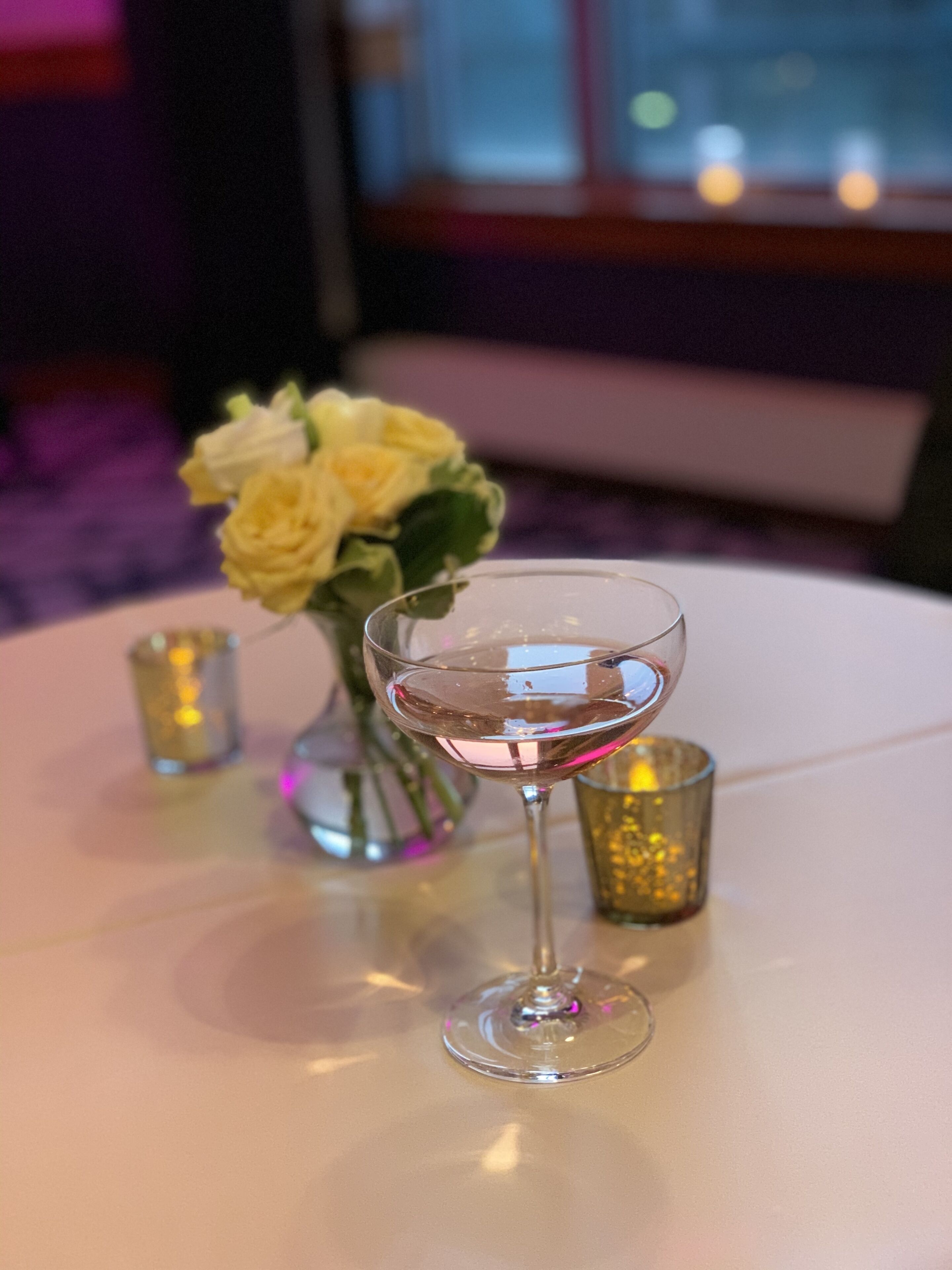 A glass of white wine on a round table with a vase of yellow roses and a small candle in the background.