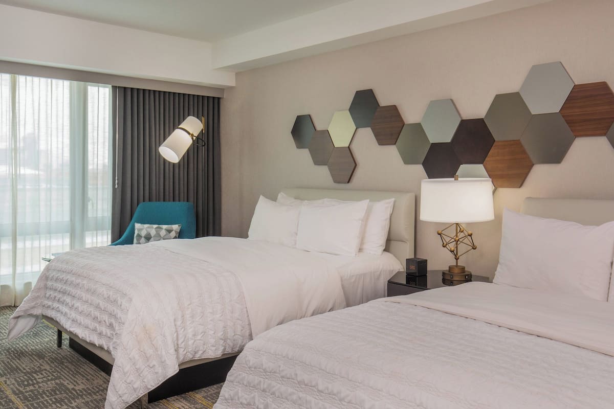 A hotel room with two beds, a large window with curtains, a wall-mounted hexagonal art piece, and a nightstand with a lamp.