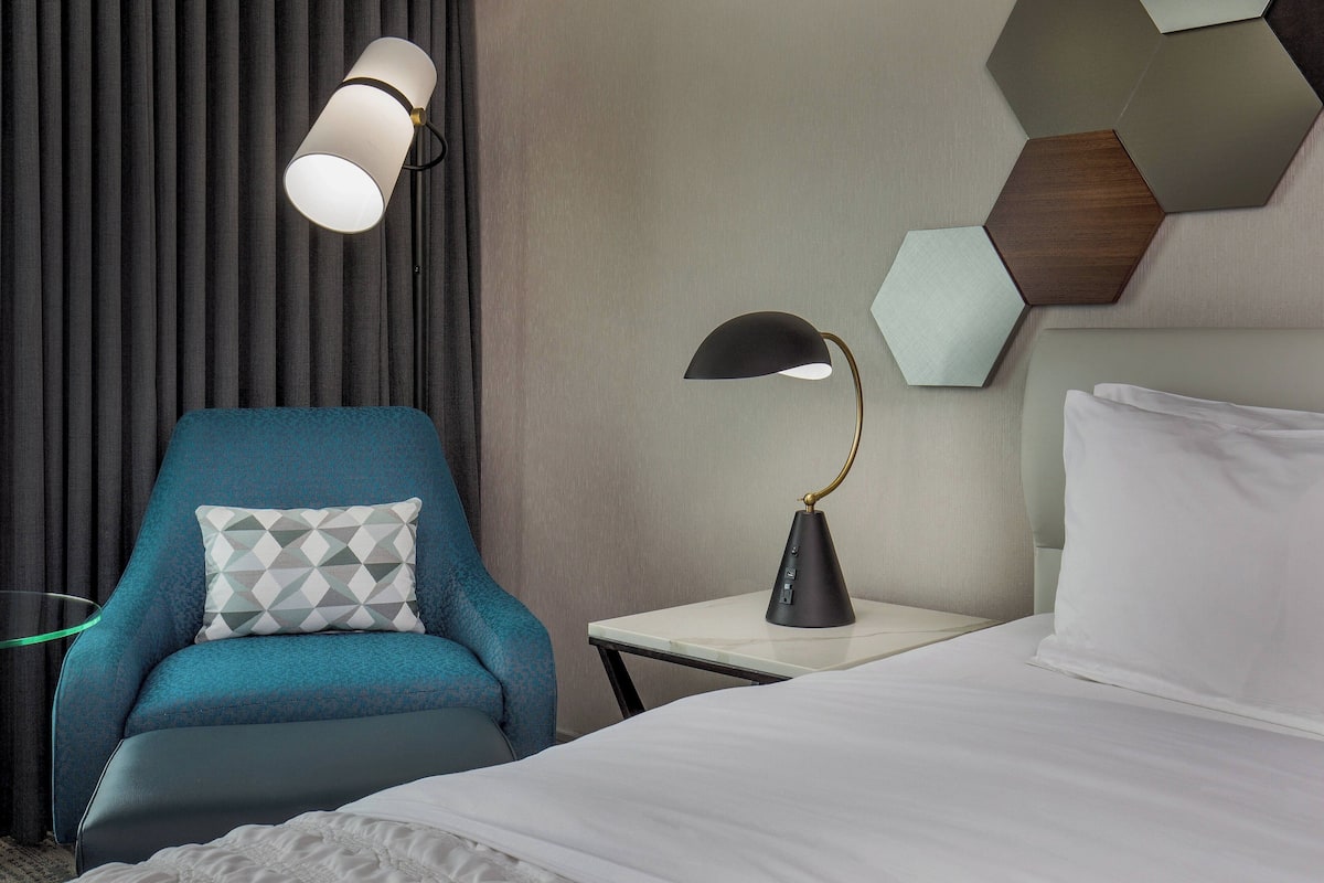 A hotel room with a bed, a chair, a nightstand, a lamp, and a wall with geometric patterns.