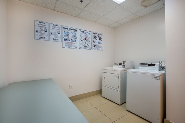 A laundry room with a bed, a washing machine, and a dryer. There are posters on the wall advertising various services.