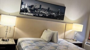 A hotel room with a bed, two bedside tables with lamps, a cityscape artwork on the wall, and a headboard. - Budgetel Inns & Suites - Atlanta Galleria Stadium (Atlanta)