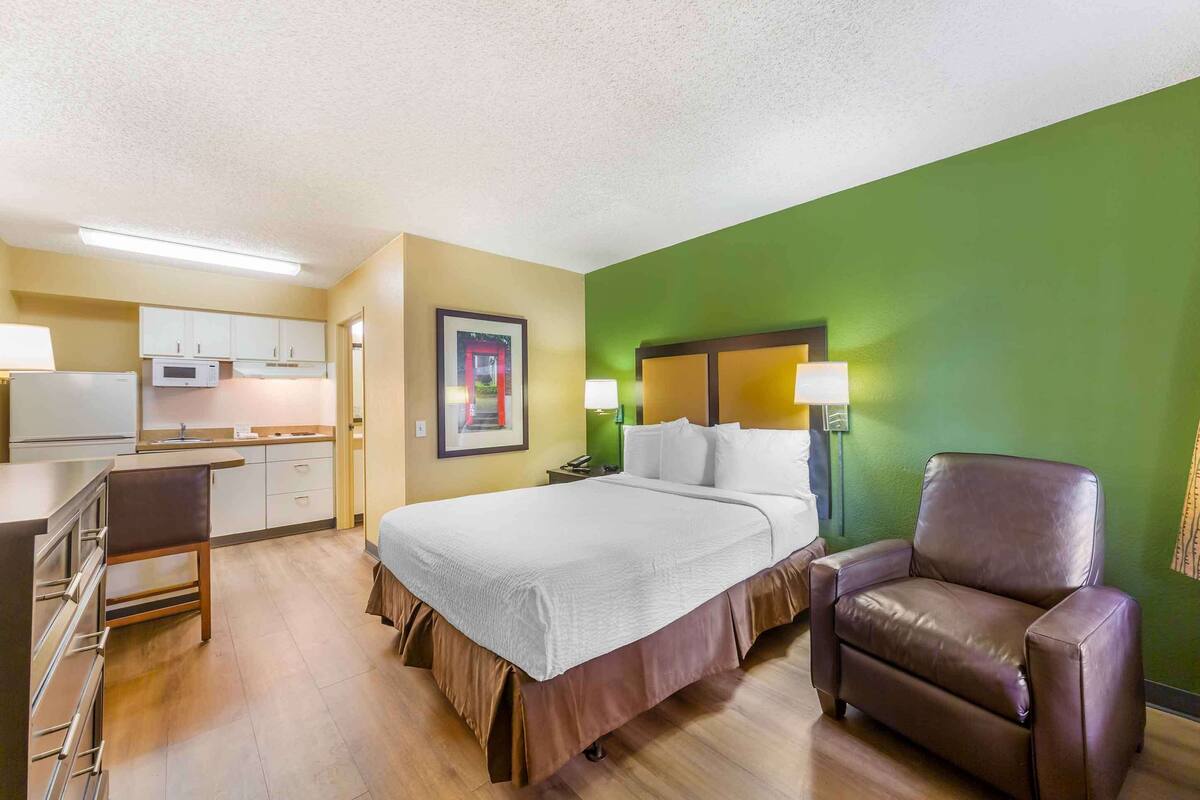 A hotel room with a bed, a desk, a chair, a microwave, and a green wall.