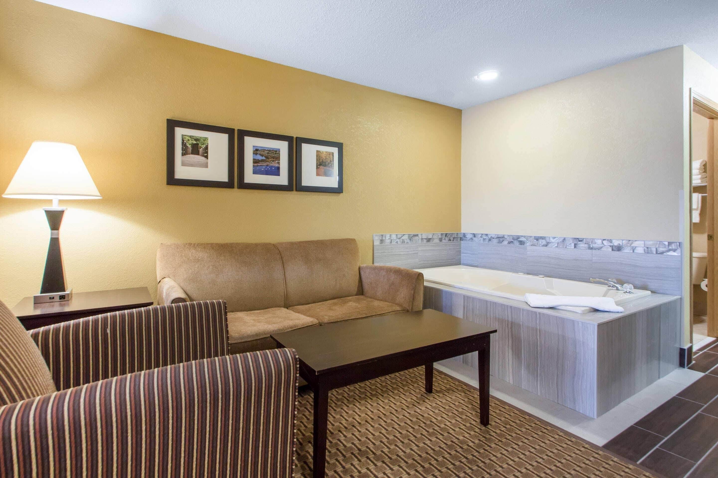 A hotel room with a sofa, armchairs, a table, a lamp, and a bathroom with a bathtub and sink.