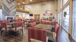 Un vaste espace dédié à la salle à manger, doté de hauts plafonds et de grandes fenêtres, comprenant plusieurs tables et des banquettes avec des sièges rembourrés de couleur rouge.