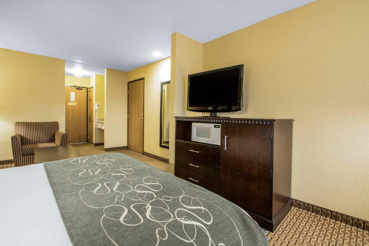 A hotel room with a bed, a television mounted on a wooden cabinet, a microwave, and a seating area with a chair.
