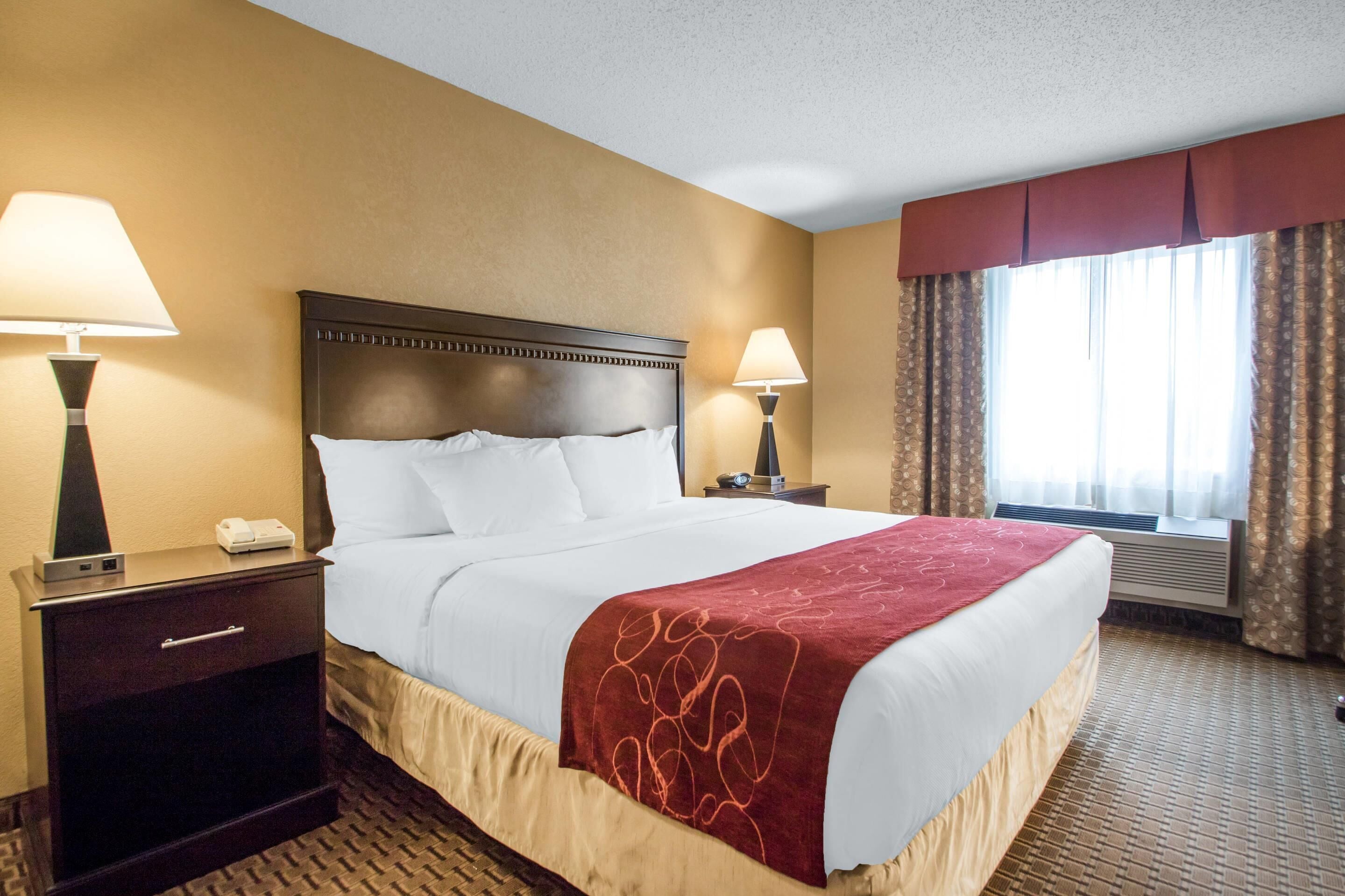 A hotel room with a large bed, two bedside tables with lamps, a window with curtains, and a wall-mounted air conditioning unit.